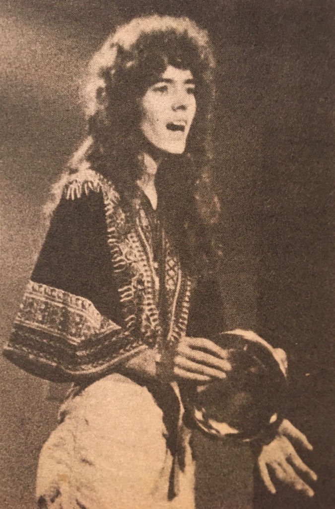 Tall thin woman with big curly hair, in old newsprint photo, singing open-mouthed holding a tambourine.